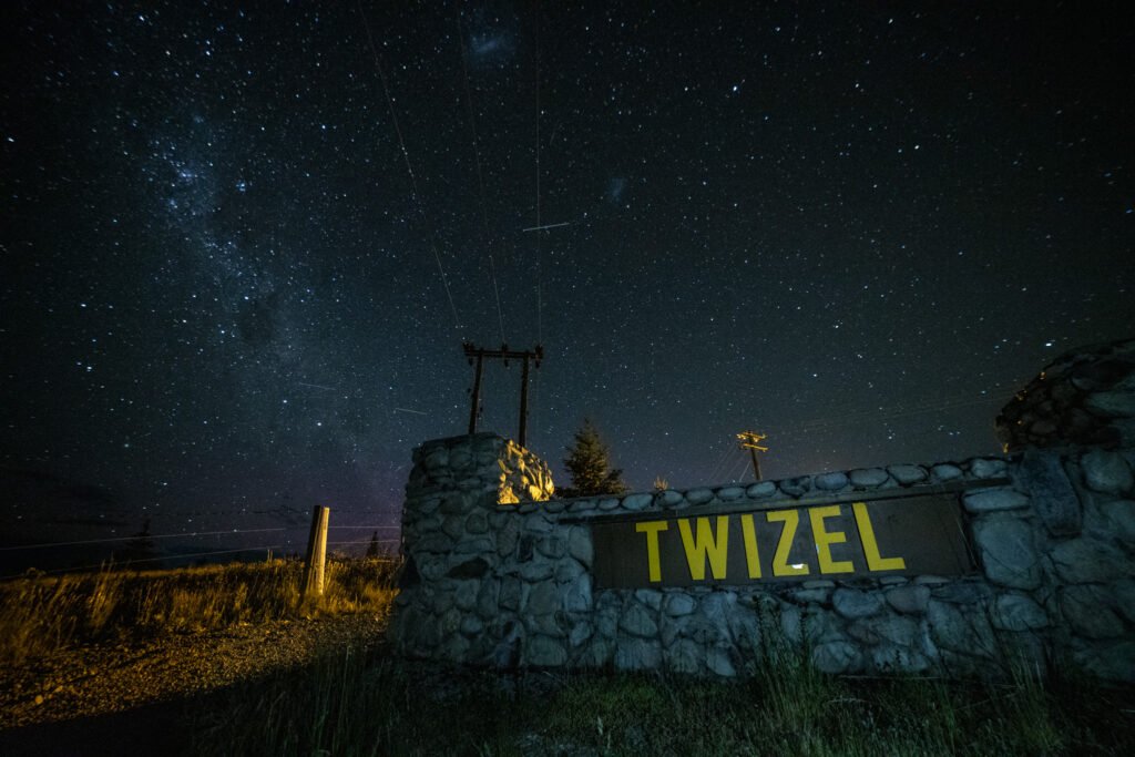 Twizel town sign illuminated under a star-filled night sky with the Southern Hemisphere constellations and a faint aurora in the background
